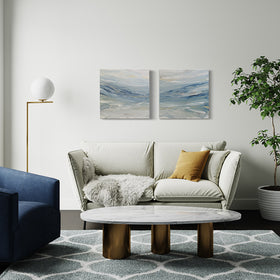A pair of abstract coastal paintings with blue, grey, white, cream, stone, teal, and yellow ochre paint by Teodora Guererra hangs on a white wall over a cream sofa. On the sofa is a cream furry throw and 2 pillows, one matching the sofa and the other yellow ochre. To the right is a large potted tree in a white pot and to the left is a table lamp with a white round glob and a blue chair which is against a window. A white marble coffee table with brass legs sits in front on a grey and white patterned rug.