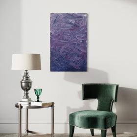 A tonal purple and pink textured abstract painting by Teodora Guererra hangs on a white wall above a green velvet chair and an end table of glass and crome with books and a table lamp of silver with a cream shade. On one of the books sits a green glass.