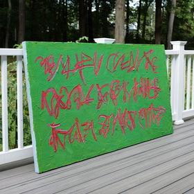 An original green canvas painting with red graffiti-style lettering, leans against a white wooden railing on a gray deck, with trees in the background.