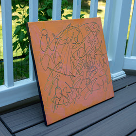 An abstract text based painting with a peach-orange background and teal carved scribbled line art, leaning against a white railing on a gray deck with green grass in the background.