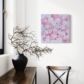 A close up of a very textured abstract painting with circle type abstract flowers in pink and white hangs on a white wall above a chair, A console sits to the left with a pot of dried decorative branches.