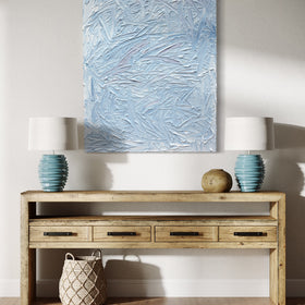 A blue and white textured painting hangs on a white wall above a natural wood credenza with two matching teal table lamps and a woven basket on the floor.