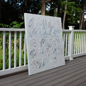 An abstract canvas text based painting with thick, textured impasto brushstrokes in shades of gray, white, and muted blue, leaning against a white wooden railing on a dark wood deck with a lush green garden background.