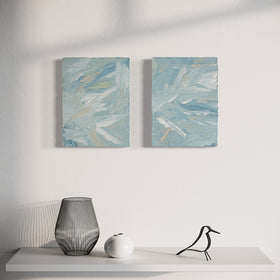 A pair of thickly painted paintings in teal, sea foam green, celadon, white and hints of orange and yellow by Teodora Guererra hang on a white wall over a white shelf with a metal bird sculpture, metal wire vase and white smaller ceramic vase sitting on it. The two painting are like wall sculptures.