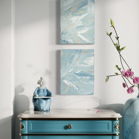 A pair of thickly painted paintings in teal, sea foam green, celadon, white and hints of orange and yellow by Teodora Guererra hang on a white wall over an ornate teal end table with white marble top. To the right is a marble vase with pink flowers and sitting on the table is a figurine of an asian woman in a teal dress. The two painting are like wall sculptures.