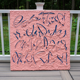 A square text based painting featuring abstract navy blue calligraphic brushstrokes over a textured pink background, displayed on a wooden deck railing with a white fence and green trees in the background.