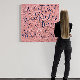 A woman with long blonde hair stands with her back to the camera, looking at a large textured pink canvas painting with dark blue calligraphic lettering mounted on a white gallery wall.
