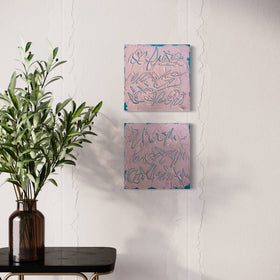Two square abstract texted based paintings with pink backgrounds and teal line work hang on a white wall above a black side table with a green plant in a brown vase.