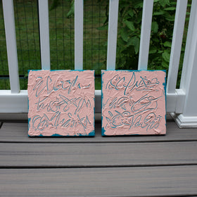 Two square pink textured abstract text based paintings with teal abstract brushstrokes, displayed side-by-side on a gray composite deck in front of a white railing with green foliage in the background.