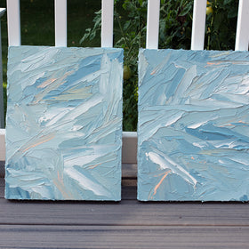A pair of thickly painted paintings in teal, sea foam green, celadon, white and hints of orange and yellow by Teodora Guererra leaning on a deck outside. The two painting are like wall sculptures.