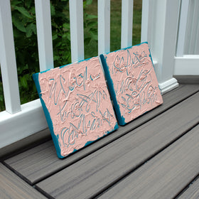Two square textured texted based paintings leaning on a gray wooden deck railing. The canvases have a dusty pink face with teal abstract brushstrokes and teal edges, with green foliage visible behind the white railing.