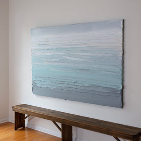 A white, teal and grey heavily textured abstract painting like sculpture by Teodora Guererra hangs on a white wall in her studio over a bench on an angle.