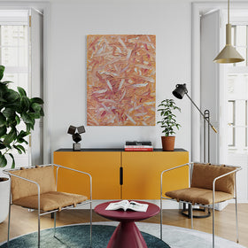 A orange, pink, coral and white thickly textured abstract painting hangs on a white wall above a mid-century yellow orange credenza with a black top. On the credenza is a sculpture, books and potted plant. In front of the credenza are 2 modern chairs with yellow ochre cushions. A low red table is in front of the chairs and sits on a grey rug.