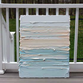 An abstract painting with heavily textured white, sherbet orange and teal blue green paint sitting on the artists deck outside.