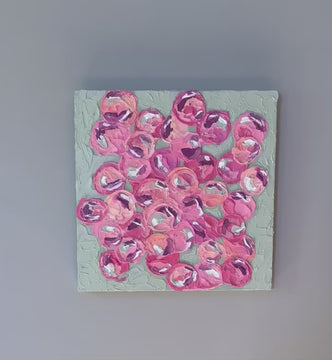 A video of a very textured abstract painting with celadon background and circle type abstract flowers in pink, peach, plum and white hangs on a white wall.