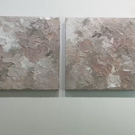 A video of a pair of thickly textured abstract paintings in light pink, grey, coral and white by Teodora Guererra hanging on a white wall.