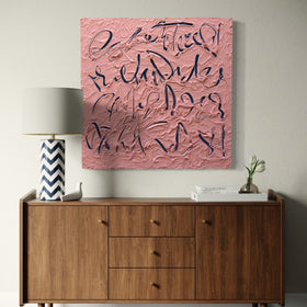 A square text based pink textured painting with dark blue abstract calligraphy hangs on a light gray wall above a mid-century modern wooden sideboard with a lamp and small plant.