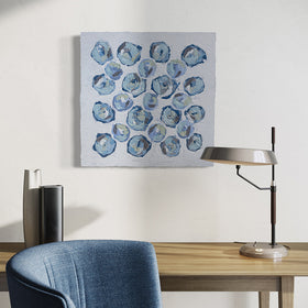 A very textured abstract painting with circle type abstract flowers in blue, teal, green, plum hangs on a white wall above a maple desk with a lamp and vases. A blue chair sits in front of it.