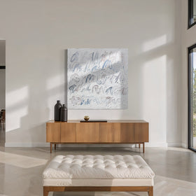 A large square abstract canvas painting with blue and gray text based calligraphic brushstrokes on a white background, displayed on a light wood console table in a modern, sunlit entryway with a tufted bench in the foreground.