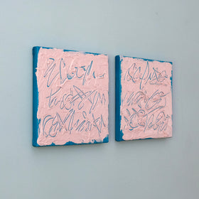 Two square text based paintings with textured pink surfaces and blue edges, featuring abstract teal line drawings, displayed side by side on a light blue-gray wall.