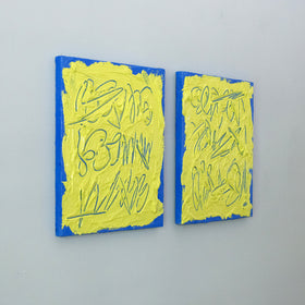 Two abstract text based painting featuring a thick, yellow-green impasto texture with blue abstract scribbled lines and a bright blue background hang on a grey wall, side view.