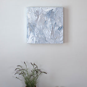 A painting is hung above a shelf. It is decorated in white, blue and grey paint applied in a thick impasto to its wooden surface. Wired and ready to hang.