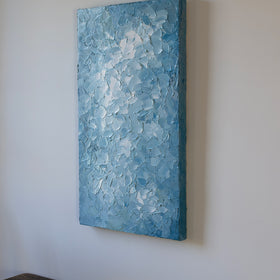 A right side-view of a blue and white abstract painting by Teodora Guererra hangs on a white wall above a dark wood bench.