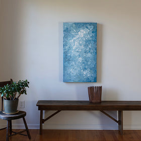 A blue and white textured abstract painting by Teodora Guererra hangs vertically on a white wall above a dark wood bench with a sculpture on the bench. A chair is to the left with a plant on it.