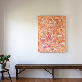 A orange, pink, coral and white thickly textured oil abstract painting hangs on a white wall in the artists studio over a bench with a yucca plat sitting to the left on a chair..